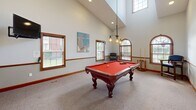 Located at 201 Eaton Ridge Dr in Northfield, Ohio, this dedicated game room is a well-appointed recreational space designed for leisure and entertainment. The room's most striking architectural feature is its dramatically vaulted ceiling, which rises to impressive heights and creates a remarkably open, airy atmosphere. Tall arched windows and multiple standard windows with wood-trimmed frames line the walls, flooding the space with generous natural light and offering views of the surrounding exterior.

At the heart of the room sits a ornately carved, full-size billiard table with a bold red felt surface, positioned as the focal centerpiece beneath a stylish multi-bulb industrial-style pendant light fixture that provides both functional and ambient illumination. Soft, neutral carpeting covers the floor throughout, adding comfort underfoot. Warm wood chair rail molding and coordinating baseboards run along the perimeter, lending a refined, classic character to the space.

The walls are adorned with vibrant contemporary artwork, including colorful abstract canvas panels that add visual energy and personality. A seating area with a cushioned chair offers a comfortable spot for spectators. A glass-paneled door provides access to an adjoining area. This game room combines architectural elegance with recreational functionality, making it an inviting and versatile space.