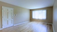 This single-floor space offers a clean and functional layout designed to maximize comfort and usability. The main living area is finished with light hardwood flooring that adds warmth and a sense of openness throughout the space. The walls are painted in a soft, warm tan tone, creating a welcoming and neutral ambiance that complements a variety of décor styles. A large window fitted with vertical blinds serves as a focal point of the room, allowing generous amounts of natural light to pour in and brighten the interior. White baseboards provide a crisp contrast against the warm walls and light floors, adding a polished finishing touch. A standard interior door leads to an additional area of the space, suggesting a practical and connected floor plan. The overall atmosphere is bright, airy, and move-in ready, offering a versatile blank canvas well-suited for a range of uses. The straightforward design prioritizes functionality while maintaining a clean aesthetic that feels both comfortable and inviting.