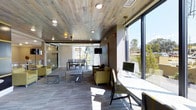 This contemporary single-floor space is defined by its thoughtful design and welcoming atmosphere. A standout feature is the expansive wood-paneled ceiling, finished in a weathered, reclaimed aesthetic that adds warmth and texture to the interior. Recessed lighting complements the natural light that pours in through large, floor-to-ceiling windows lining one side of the space, offering open views of the surrounding greenery and blue skies beyond.

The layout is organized into distinct functional zones that flow seamlessly into one another. A comfortable lounge area features olive-toned upholstered seating arranged around low-profile coffee tables, creating an inviting spot for casual gatherings or relaxed conversation. Adjacent to this is a dedicated workstation area, equipped with desktop computers and a sleek long desk positioned directly beside the expansive windows—making it an ideal environment for focused productivity.

Further into the space, a dining area with a full table and chairs provides a setting suited for meetings or communal meals, with access to additional natural light through sliding glass doors. The flooring throughout consists of patterned carpet in cool, neutral tones, tying the various areas together cohesively. The overall ambiance strikes a balance between modern functionality and comfortable design.