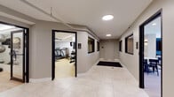 This single-floor amenity space is thoughtfully designed to offer a comprehensive range of recreational and social facilities. The layout flows seamlessly from one area to the next, connecting distinct zones that cater to fitness, relaxation, and entertainment.

The fitness center is generously sized and outfitted with a wide array of cardio and strength-training equipment, including treadmills, weight machines, and free weights, all set on light hardwood-style flooring beneath bright overhead lighting. An adjacent hallway with large windows and tile flooring provides easy access to the gym and nearby facilities.

The lounge and entertainment area features rich blue textured accent walls, a large wall-mounted flat-screen television flanked by sleek vertical LED light panels, and plush neutral seating arranged around a dark marble coffee table — creating an inviting, sophisticated atmosphere. This space connects directly to a game room equipped with a foosball table, a billiards table, and additional seating.

The kitchenette is a design highlight, showcasing warm wood cabinetry, white marble-veined countertops and backsplash, light hardwood flooring, and striking vertical wood slat wall paneling that extends to a slatted wood ceiling. Dramatic black and gold pendant lights complete the upscale aesthetic. A casual dining nook with high-top tables and navy blue bar stools provides a relaxed setting for socializing.