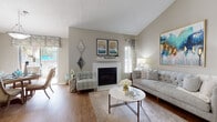 Located in Peachtree Corners, Georgia, this single-story residence offers a well-planned layout with distinct living spaces connected by a tastefully decorated hallway. The entry corridor features hardwood-style flooring and wall art, setting a welcoming tone throughout.

The living room is a standout space, characterized by striking vaulted ceilings that lend an open, airy feel. A white-mantled fireplace serves as the room's focal point, flanked by windows that invite natural light. Hardwood-style flooring continues here, complementing the neutral wall tones.

The dining area is positioned near sliding glass doors that open to an outdoor space, offering views of greenery and a patio area beyond. A pendant light fixture anchors the space above a round dining table.

The kitchen features white cabinetry with brushed hardware, stone-look countertops, and stainless steel appliances, including a range, microwave, and dishwasher, alongside light hardwood-style flooring.

Both bedrooms are carpeted and receive natural light through windows with decorative drapery. The primary bedroom includes ample space for a dresser and seating area. The bathroom presents stone-look countertops, white cabinetry, and a large vanity mirror.