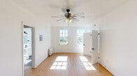 This single-floor space is designed with a clean, minimalist aesthetic that emphasizes light, openness, and functional simplicity. Throughout the unit, warm-toned hardwood flooring creates a cohesive and inviting foundation, complementing the crisp white walls that reflect natural light throughout.

The main living area is a highlight of the space, featuring two classic multi-pane windows that frame views of the surrounding urban neighborhood. Sunlight streams generously through these windows, brightening the room at various times of day. A ceiling fan with an integrated light fixture anchors the ceiling, providing both airflow and ambient lighting. A traditional radiator along the wall speaks to the building's character, while a wall-mounted intercom panel adds a practical touch.

The floor plan, visible from the doll's-eye view, reveals a compact but thoughtfully arranged layout that includes a separate room accessible through white panel doors, and a dedicated storage area outfitted with a full-width shelving rod — ideal for organized closet use.

The bathroom stands out with floor-to-ceiling white subway tile accented by dark grout and matte black fixtures, including a sleek tub spout and shower head. A window above the tub allows natural light to fill the space while offering an elevated view of the cityscape, making it both functional and visually striking.