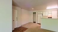 This single-floor space offers a straightforward and functional layout designed for comfortable everyday living. The main living area is finished with wall-to-wall carpeting that adds warmth underfoot, while the entryway features durable hardwood-style flooring that provides a clean transition into the space. Recessed lighting in the ceiling delivers consistent illumination throughout the room, and neutral white walls keep the interior feeling bright and open.

A generously sized walk-in closet off the main living area provides ample storage, fitted with an interior rod for hanging items. The entry door is equipped with multiple locks, offering added security and peace of mind.

The kitchen is positioned adjacent to the living space and is separated by a partial countertop bar that creates a natural division between the two areas. White upper cabinetry lines the kitchen wall, offering practical storage, and a full-size white refrigerator anchors the space. The kitchen's tile or hard-surface flooring contrasts neatly with the carpeted living area, making it easy to maintain.

Overall, the space presents a clean, move-in-ready environment with a simple and efficient design, well-suited for those seeking a low-maintenance, functional living arrangement.