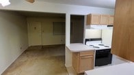 This single-floor space features an open layout that connects the kitchen and living areas, creating a cohesive and functional environment. The kitchen is equipped with a white coil-burner range and oven, light oak cabinetry providing ample storage, and an under-cabinet range hood. A peninsula-style countertop extends into the space, offering additional workspace and a natural division between the kitchen and the adjoining living area. The cabinetry features a warm wood tone that complements the neutral color palette throughout. The living area is carpeted and receives modest natural light, with a textured ceiling adding subtle visual detail overhead. A doorway visible from the main space leads to an additional room or hallway, suggesting a multi-room floor plan. The front entry door is visible from the living area, contributing to the open sightlines across the unit. The overall aesthetic is straightforward and utilitarian, with light-toned walls and neutral finishes throughout. The layout prioritizes practicality and ease of movement, making efficient use of the available space on this single level.