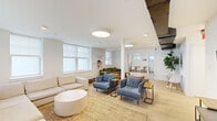 This well-appointed single-floor space offers a cohesive open layout that seamlessly connects its key areas, finished throughout with light hardwood flooring and a crisp, white palette that enhances the sense of openness and warmth.

The expansive living room is a standout feature, divided into two distinct seating zones. One side is anchored by a large L-shaped cream sectional sofa paired with a round stone coffee table atop a natural woven rug, while the other zone offers a green velvet tufted sofa, cognac leather armchair, and rich wooden accent tables on a patterned rug. Multiple large windows flood both areas with natural light, and a wall-mounted flat-screen TV with a media console rounds out the space.

The combined dining room and kitchen area is both functional and stylish. The kitchen features white shaker cabinetry, subway tile backsplash, stainless steel appliances including a gas range and French door refrigerator, and a large white island with tan leather counter stools beneath matte gray pendant lights. Adjacent to the kitchen, a long solid oak dining table with modern white chairs sits beneath a row of windows adorned with framed artwork, creating an inviting communal dining experience.

A connecting hallway ties the layout together, contributing to the space's efficient and intuitive flow throughout.