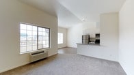 This single-floor space showcases an open-concept layout that seamlessly connects the living area to the kitchen, creating a functional and airy environment. The living area is finished with soft, neutral-toned carpet, providing a comfortable and inviting atmosphere. Large gridded windows along the exterior wall allow generous natural light to fill the space, offering views of the surrounding neighborhood.

The vaulted ceiling adds a sense of vertical openness, enhancing the overall spaciousness of the unit. A wall-mounted HVAC unit provides convenient climate control directly in the living area.

The kitchen is positioned on an elevated counter peninsula, offering a natural division between the cooking and living spaces. It features white cabinetry, dark countertops, and a full suite of stainless steel appliances, including a refrigerator, microwave, and range. A built-in sink completes the practical and clean kitchen setup.

The interior palette is kept consistently neutral throughout, with white walls and light finishes that contribute to a clean, move-in-ready aesthetic. The combination of natural light, vaulted ceilings, and an open floor plan makes this space feel welcoming and versatile.