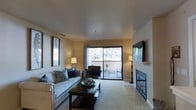 This single-story living space centers around a well-appointed living room that balances comfort and functionality. The room is finished with neutral-toned walls and smooth ceilings, creating a warm and inviting atmosphere throughout. Soft carpeting anchors the main seating area, while tile flooring near the fireplace adds a practical and visually distinct transition.

A standout feature of the living room is the fireplace, framed by a clean white mantel and set against tile surround, serving as a natural focal point on one wall. Opposite, a wall-mounted flat-screen television complements the entertainment setup. A large sliding glass door floods the space with natural light and provides direct access to an outdoor patio area, offering pleasant views of surrounding greenery and trees.

The living area is generously sized, comfortably accommodating a full sofa with accent pillows, a dark wood coffee table, and an additional seating chair near the patio door. Warm ambient lighting from floor and table lamps enhances the cozy feel during evening hours. The overall design aesthetic is understated and practical, making it a functional and welcoming living environment.