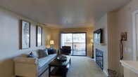 This single-story living space centers around a well-appointed living room that balances comfort and functionality. The room is finished with neutral-toned walls and smooth ceilings, creating a warm and inviting atmosphere throughout. Soft carpeting anchors the main seating area, while tile flooring near the fireplace adds a practical and visually distinct transition.

A standout feature of the living room is the fireplace, framed by a clean white mantel and set against tile surround, serving as a natural focal point on one wall. Opposite, a wall-mounted flat-screen television complements the entertainment setup. A large sliding glass door floods the space with natural light and provides direct access to an outdoor patio area, offering pleasant views of surrounding greenery and trees.

The living area is generously sized, comfortably accommodating a full sofa with accent pillows, a dark wood coffee table, and an additional seating chair near the patio door. Warm ambient lighting from floor and table lamps enhances the cozy feel during evening hours. The overall design aesthetic is understated and practical, making it a functional and welcoming living environment.
