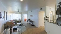 This single-floor residential unit offers a comfortable and functional layout designed for everyday living. The main living area is generously proportioned, featuring warm carpet flooring that extends throughout the space, creating a cozy and inviting atmosphere. Large windows dressed with sheer and accent curtains allow abundant natural light to fill the room, enhancing the open feel of the interior. A ceiling fan is centrally positioned in the living area, providing both practical airflow and a subtle decorative element. The smooth, textured ceilings add a classic finish to the space. A distinctive arched doorway serves as an architectural highlight, elegantly framing the transition between the living area and the adjoining hallway, which leads to additional rooms. The neutral wall tones in soft gray-blue hues create a calm and versatile backdrop throughout. A dedicated entryway or transitional space is visible near the kitchen pass-through area, hinted at by a raised countertop edge, suggesting a connected kitchen layout. The hallway provides access to private areas of the unit, with a clean, white-door finish maintaining a bright and cohesive interior aesthetic. Overall, the space combines practical design with welcoming details suited for comfortable residential living.