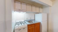 This single-floor unit offers a practical and straightforward layout designed to maximize usable space. The kitchen serves as a central feature, equipped with a gas range, a stainless steel sink, warm-toned wood cabinetry, upper cabinets for ample storage, a white refrigerator, and a tiled backsplash. A peninsula with a speckled countertop extends outward, creating a natural breakfast bar that separates the kitchen from the adjacent living area and offering additional prep and serving space.

The living area is generously sized, finished with wall-to-wall carpeting in a neutral beige tone that carries throughout the unit. White walls and a clean ceiling keep the space feeling bright and open. A wall-mounted thermostat/control panel is conveniently positioned near the entry corridor. A separate bedroom area benefits from a wall-mounted air conditioning unit and a horizontal window that allows natural light to filter in.

Storage is well-addressed through a spacious walk-in closet fitted with a full-length hanging rod and carpeted flooring, in addition to a standard reach-in closet accessible from the hallway. The bathroom features a full soaking tub and shower combination, with crisp white square tiles lining the walls and a bold black-and-white checkered tile border adding a decorative accent. The overall ambiance is clean, neutral, and move-in ready.