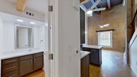 This two-floor loft-style space masterfully combines industrial heritage with contemporary design. Exposed brick walls serve as a defining architectural feature throughout, lending warmth and texture that contrast beautifully with crisp white walls and smooth hardwood flooring found across both levels. Soaring ceilings amplify the sense of openness, while ceiling fans provide both function and a subtle industrial flair.

Natural light flows generously through large windows fitted with horizontal blinds, illuminating the generous living areas and highlighting the rich tones of the hardwood floors. A staircase with metal railings connects the two floors, visible in the overall layout of the space.

The kitchen is well-equipped and thoughtfully designed, featuring dark cabinetry with modern hardware, light countertops, a double sink, and black appliances including a range, dishwasher, and refrigerator. Ample upper and lower cabinet storage makes this a highly functional cooking space.

The bathroom offers a clean, modern aesthetic with a combination tub and shower outfitted with accessibility grab bars, a vanity with dark cabinetry, a framed mirror, and bright recessed lighting overhead.

Throughout the space, the interplay of raw brick, warm wood tones, and contemporary finishes creates a distinctive ambiance that is both urban and inviting.
