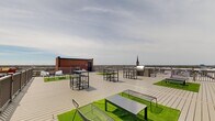Perched atop a building in the heart of downtown Toledo, Ohio, this rooftop terrace offers an expansive open-air retreat with sweeping panoramic views of the city skyline and surrounding urban landscape. The generously sized composite wood deck provides a versatile outdoor gathering space, thoughtfully appointed with multiple high-top table-and-chair sets ideal for socializing and dining al fresco. Interspersed throughout the deck are patches of vibrant artificial turf, each paired with modern low-profile benches and tables, creating distinct lounge areas that add a touch of greenery to the urban setting. A dedicated grilling station invites outdoor cooking and entertaining. The terrace is enclosed by sturdy perimeter railings that ensure safety while preserving unobstructed sightlines across the Toledo cityscape, including a prominent church steeple and distant bridges visible on the horizon. The building's original brick parapet walls frame portions of the space, lending an industrial character that complements the contemporary furnishings. Open to the sky, the terrace benefits from abundant natural light throughout the day, making it a dynamic space well-suited for both casual relaxation and larger social gatherings in the heart of Toledo.