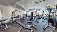 This fitness center is a fully outfitted, single-floor facility designed to support a wide range of workout routines. The space features soaring vaulted ceilings that lend an open, airy feel, enhanced by large windows along the walls that invite abundant natural light throughout the day. Large ceiling fans circulate air across the floor, contributing to a comfortable workout environment alongside recessed lighting overhead.

The cardio zone is well-stocked with multiple treadmills, stationary bikes, and additional cardio machines, all positioned beneath wall-mounted flat-screen televisions. A full mirrored wall runs along one side of the room, visually expanding the space and providing users with form-checking capability during exercise.

The strength training area is equally comprehensive, housing a Smith machine, cable crossover station, multi-functional cable machines, a selection of benches, and a dedicated free weights section with a full rack of dumbbells and kettlebells in graduated sizes. Barbells and weight plates are neatly organized on storage racks throughout the space.

The flooring throughout is a durable dark rubber material suited for heavy equipment and high-traffic use. The clean, neutral color palette of the walls paired with the abundance of natural light creates a focused yet welcoming atmosphere, making this fitness center a functional and well-designed space for daily training.
