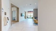 Located at 100 Boucle Jeanne Circle in Maitland, Florida, this thoughtfully designed single-floor amenity space offers a generous, open layout that seamlessly blends comfort, functionality, and modern aesthetics. Large-format white tile and light wood flooring flow throughout, creating a clean, cohesive foundation across the various zones.

The welcoming lounge area is a standout feature, anchored by a bold geometric accent wall in teal and gold tones. Stylish seating — including sculptural gold woven armchairs and a tufted gray sofa — is arranged around a patterned area rug, creating an inviting gathering space. Nearby, a separate seating nook features striped upholstered chairs, wall-mounted sconces, and decorative circular mirrors framed in warm wood, all positioned beside a wall-mounted flat-screen display.

Floor-to-ceiling windows and arched glass panels flood the interior with natural light and offer views of the surrounding Florida landscape, including palm trees and open skies. A long communal work table with modern leather chairs sits beneath a dramatic rope-hung pendant fixture, providing a stylish co-working area.

The kitchen is appointed with elegant marble-look stone countertops, open wood shelving, and warm cabinetry. Glass-paneled private office spaces and art-adorned hallways connect the various areas, rounding out a space designed for both productivity and community.