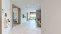 Located at 100 Boucle Jeanne Circle in Maitland, Florida, this thoughtfully designed single-floor amenity space offers a generous, open layout that seamlessly blends comfort, functionality, and modern aesthetics. Large-format white tile and light wood flooring flow throughout, creating a clean, cohesive foundation across the various zones.

The welcoming lounge area is a standout feature, anchored by a bold geometric accent wall in teal and gold tones. Stylish seating — including sculptural gold woven armchairs and a tufted gray sofa — is arranged around a patterned area rug, creating an inviting gathering space. Nearby, a separate seating nook features striped upholstered chairs, wall-mounted sconces, and decorative circular mirrors framed in warm wood, all positioned beside a wall-mounted flat-screen display.

Floor-to-ceiling windows and arched glass panels flood the interior with natural light and offer views of the surrounding Florida landscape, including palm trees and open skies. A long communal work table with modern leather chairs sits beneath a dramatic rope-hung pendant fixture, providing a stylish co-working area.

The kitchen is appointed with elegant marble-look stone countertops, open wood shelving, and warm cabinetry. Glass-paneled private office spaces and art-adorned hallways connect the various areas, rounding out a space designed for both productivity and community.