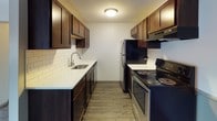 This single-floor residence offers a well-organized layout with distinct living spaces designed for everyday comfort and functionality. The open living area features dark hardwood-style flooring, a ceiling fan, and a window allowing natural light, while a louvered closet door provides practical storage near the entryway. The space flows naturally toward the kitchen, which is outfitted in a galley configuration with rich dark wood cabinetry lining both walls, white subway tile backsplash, light countertops, a single-basin sink, and stainless steel appliances including a refrigerator and electric range. The bedrooms are carpeted and each equipped with a ceiling fan and window blinds. The larger bedroom benefits from dual windows that invite generous natural light, while the secondary bedroom features two-tone wainscoting along the lower walls. Both rooms include baseboard heating. The bathroom is clean and functional, presenting white square mosaic tile flooring, a full-size tub and shower combination, a toilet, and a vanity with tiled backsplash and dark wood cabinetry. Throughout the residence, a consistent neutral color palette and practical finishes create a cohesive and move-in ready environment.