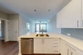 This single-floor residence is designed with a clean, contemporary aesthetic throughout. The open-concept kitchen serves as the focal point of the living area, featuring sleek white cabinetry, light quartz countertops, and a subway tile backsplash in a soft grey-blue tone. A peninsula with an undermount sink and pendant lighting above creates a natural division between the kitchen and the living area beyond. Stainless steel appliances, including a refrigerator, range, built-in microwave, and dishwasher, complete the well-equipped cooking space. In-unit stacked washer and dryer are conveniently tucked into a dedicated closet nearby. Wood-style flooring runs through the main living areas, contributing to a warm, cohesive feel, while large windows allow natural light to fill the space and offer outdoor views. The bedroom features soft carpeting and a generously sized window with blinds. An adjacent bathroom is outfitted with a wide vanity, quartz countertop, white cabinetry, and a large mirror. A walk-in closet with built-in shelving and hanging rods provides ample organized storage. The overall palette of soft neutrals and whites gives the space a bright, move-in-ready character.