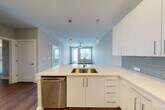 This single-floor residence is designed with a clean, contemporary aesthetic throughout. The open-concept kitchen serves as the focal point of the living area, featuring sleek white cabinetry, light quartz countertops, and a subway tile backsplash in a soft grey-blue tone. A peninsula with an undermount sink and pendant lighting above creates a natural division between the kitchen and the living area beyond. Stainless steel appliances, including a refrigerator, range, built-in microwave, and dishwasher, complete the well-equipped cooking space. In-unit stacked washer and dryer are conveniently tucked into a dedicated closet nearby. Wood-style flooring runs through the main living areas, contributing to a warm, cohesive feel, while large windows allow natural light to fill the space and offer outdoor views. The bedroom features soft carpeting and a generously sized window with blinds. An adjacent bathroom is outfitted with a wide vanity, quartz countertop, white cabinetry, and a large mirror. A walk-in closet with built-in shelving and hanging rods provides ample organized storage. The overall palette of soft neutrals and whites gives the space a bright, move-in-ready character.
