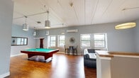 Located at 1317 East St in New Britain, CT, this single-floor community lounge is a well-appointed recreational space designed for comfort and social engagement. The open-plan layout is anchored by rich hardwood flooring and accented by cool gray walls, creating a clean, contemporary aesthetic throughout.

The main living area is generously sized, featuring a large black leather sectional sofa and matching accent chairs arranged around a wooden coffee table, all centered beneath a modern circular pendant light. A wall-mounted flat-screen TV serves as a focal point for the lounge seating zone. Multiple windows along the perimeter admit ample natural light and offer views of the surrounding area, enhancing the open and airy feel of the space.

A dedicated game zone features a full-size pool table positioned beneath a row of sleek, cylindrical pendant lights suspended from the high ceiling — a standout architectural detail that adds volume and character to the room. Nearby, a high-top table with bar-height stools provides an additional casual seating option.

Practical amenities include a beverage vending machine and a snack vending machine, conveniently accessible within the space. A counter area and closet offer additional functional storage. A wall-mounted mini-split unit ensures year-round climate comfort. Together, these features make this lounge an inviting, fully equipped communal retreat.