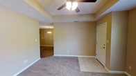 This single-floor residence offers a thoughtfully designed layout that balances comfort and functionality. The living area serves as the heart of the home, distinguished by a tray ceiling that adds architectural depth and dimension to the space. A ceiling fan with integrated lighting is centrally mounted, providing both airflow and ambient illumination. The room features plush wall-to-wall carpeting that transitions seamlessly into tile flooring near the entryway, creating a natural delineation between spaces. Warm, neutral-toned walls lend a welcoming atmosphere throughout. A solid entry door with sidelights provides a secure and classic point of entry. Just beyond the living area, an adjoining room — likely a dining space — is visible through a wide doorway, accented by a decorative chandelier that adds a touch of elegance. The open sightlines between rooms enhance the sense of spaciousness and allow for a natural flow through the home. White baseboards provide clean contrast against the neutral walls, contributing to a tidy, well-maintained aesthetic. Overall, this space presents a cozy yet refined interior with classic residential finishes throughout.