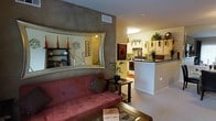 This single-floor space showcases a warm and eclectic interior design with a seamless open-concept layout. The living area is a standout, featuring a striking curved accent wall finished in a textured gray plaster, which frames a large decorative mirror and built-in display shelving. These shelves are adorned with decorative objects, adding character and depth to the space. A plush red sofa anchors the seating area, complemented by a glass-top coffee table and an abundance of indoor plants that bring a natural, lively energy to the room. The transition to the kitchen is fluid and unobstructed, with a raised breakfast bar serving as a functional divider between the two areas. The kitchen is equipped with white cabinetry, modern appliances including a range and overhead microwave, and a dark countertop that provides visual contrast. Warm overhead lighting illuminates the kitchen, while natural and ambient light fills the living space. The overall ambiance is cozy and inviting, blending earthy tones with warm lighting to create a space that feels both stylish and livable. Decorative artwork and thoughtful accessory placement throughout add a personalized, curated quality to the interior.