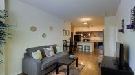 This single-floor residence showcases an open-concept layout that connects the living area and kitchen in one cohesive, flowing space. The living room is anchored by a comfortable gray sofa adorned with decorative throw pillows, positioned atop a floral-patterned area rug over warm hardwood flooring that extends throughout the unit. A simple black coffee table completes the seating arrangement, while decorative metallic wall plates add an artistic touch to the neutral walls.

Transitioning naturally from the living area, a compact dining space features a dark bistro-style table with matching chairs, providing an intimate setting for meals. The kitchen beyond is well-appointed with dark cabinetry, a peninsula-style breakfast bar with bar stools, and pendant lighting that casts a warm glow over the workspace. A black refrigerator and integrated appliances complete the functional kitchen setup. Track lighting and a central overhead fixture ensure ample illumination throughout the cooking and dining areas. The overall ambiance is warm and inviting, with a neutral color palette, consistent flooring, and thoughtful lighting creating a cohesive and functional living environment.
