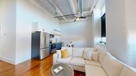 This thoughtfully designed loft-style unit blends industrial character with modern finishes across a single floor. The open-concept kitchen and living area serves as the heart of the space, where soaring ceilings with exposed ductwork and wooden beams create a striking visual backdrop. A ceiling fan anchors the airy volume above. The kitchen is outfitted with bold blue lower cabinetry, white upper cabinets, open wood shelving, stainless steel appliances, a black matte faucet, and a marble-topped island with seating — ideal for casual dining. Rich hardwood floors flow throughout, complementing the whitewashed brick walls visible in the living area, which is bathed in natural light from large industrial-style windows. A long, clean hallway connects the four bedrooms, each featuring the same warm hardwood floors and high ceilings. The primary bedroom offers generous proportions with tall walls and ample space. The shared bathroom is finished with bold black hexagonal tile flooring, large-format white wall tiles, and a glass-enclosed walk-in shower with matte black hardware. A dedicated laundry room with matching hexagonal tile provides added convenience. The overall ambiance is modern, urban, and welcoming.