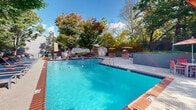This inviting outdoor pool area in Memphis, Tennessee offers a well-designed recreational retreat ideal for relaxation and leisure. The generously sized swimming pool showcases vivid turquoise water, framed by classic brick coping and blue tile accents that lend a polished, resort-like aesthetic. Depth markers are clearly visible, and lane dividers are in place to accommodate both casual swimmers and lap enthusiasts.

The surrounding pool deck provides ample space for lounging, lined with rows of blue-and-orange chaise lounges accompanied by coordinating side tables — offering a cohesive and vibrant color scheme throughout. A raised concrete patio area to one side features multiple high-top tables with red chairs and shade umbrellas, creating a comfortable spot for outdoor dining or socializing. A utility shed is conveniently located nearby for storage and equipment access.

The entire area is enclosed by a privacy fence and accented by a mature canopy of deciduous trees, delivering natural shade and a serene, park-like atmosphere. The lush greenery surrounding the space provides a tranquil backdrop that enhances the open-air environment. Under clear blue skies, this outdoor pool area delivers a bright, welcoming ambiance well-suited for community gatherings and warm-weather enjoyment.