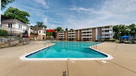 Located at 4085 Warner Ave in Hyattsville, Maryland, this outdoor amenity area serves as a central recreational hub for the surrounding multi-story residential complex. The centerpiece is a generously sized swimming pool with clear turquoise water, featuring entry stairs with handrails and clearly marked depth indicators. The pool's distinctive shape provides ample room for both lap swimming and leisure use.

Surrounding the pool is a broad, open concrete sundeck that offers plenty of space for relaxation and movement. The deck is furnished with dark-framed lounge chairs and shaded by vibrant blue umbrellas positioned throughout the area, creating comfortable spots for outdoor leisure. Additional seating areas are found near decorative stone retaining walls that add a natural, textured element to the landscape.

The complex's multi-level brick residential buildings frame the pool area on multiple sides, each featuring private balconies that overlook the amenity space. Mature trees line the perimeter, providing natural shade and greenery that softens the hardscape environment. A gated access point leads to an adjacent open lawn area, extending the outdoor recreational space further. Exterior wall-mounted lighting and tall pole lights ensure the area is well-illuminated. The overall setting is open and community-oriented, designed to encourage outdoor activity and social gathering in a well-maintained environment.