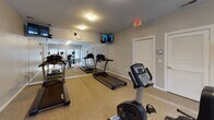 This single-floor amenity space is thoughtfully divided into two distinct functional areas: a fitness center and a private screening room. The fitness center is bright and well-lit, benefiting from natural light streaming through several windows along one wall. Large floor-to-ceiling mirrors line one side of the room, amplifying the sense of space and allowing users to monitor their form. The carpeted floor cushions a varied selection of cardio equipment, including treadmills, elliptical trainers, and stationary bikes, while wall-mounted monitors provide entertainment during workouts. An exit door provides direct outdoor access, and restroom facilities are conveniently located within the space.

Adjacent to the fitness area, the screening room delivers an intimate, cinema-like experience. Rows of rich brown leather reclining theater seats, each equipped with built-in cup holders, are arranged to face a large display screen mounted on a neutral gray wall. Elegant wall sconces with brass accents provide warm, ambient lighting, complementing the room's dedicated audio-visual setup. A raised counter area at the rear offers additional utility. Together, both areas provide a comprehensive and comfortable amenity suite.