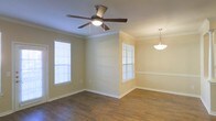 This single-story space showcases a thoughtfully designed open layout with classic architectural details throughout. The living and dining areas are defined by rich, dark hardwood flooring that adds warmth and depth to the interior. Crown molding runs along the upper perimeter of the walls, lending an elegant, finished look to the space.

Natural light flows in generously through multiple windows fitted with white horizontal blinds, brightening the warm beige-toned walls. A ceiling fan with a built-in light fixture is centrally positioned in the living area, offering both comfort and functionality. The adjacent dining area is distinguished by a stylish pendant light that hangs gracefully from the ceiling, creating a welcoming atmosphere for dining and entertaining.

A partial dividing wall separates the two areas while maintaining an open, airy feel between them. White baseboards provide clean contrast against the flooring, complementing the neutral wall tones. The overall ambiance is comfortable and inviting, combining practical design elements with classic detailing that suits a variety of interior styles.