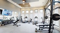 Located in Midlothian, Texas, this fitness center offers a thoughtfully designed workout space spread across a single open floor. The room makes a strong architectural impression with soaring walls, a row of clerestory windows that line the upper perimeter, and a distinctive reclaimed wood ceiling accent panel that adds a rustic, modern touch above the main exercise area. A large glass garage-style door on one wall floods the space with natural light and provides open views of the surrounding community.

The interior features durable speckled rubber flooring throughout, ideal for high-impact activity. Full-length mirrors are strategically positioned along one wall to maximize the sense of space and support proper form during workouts. Cardio equipment is well-represented, with multiple treadmills, ellipticals, a stationary bike, and a rowing machine arranged along the windowed wall. A multi-function cable machine anchors the strength training area, complemented by a rack of dumbbells, medicine balls, stability balls, and adjustable benches for versatile training options. A large flat-screen television is mounted prominently, providing an engaging visual focal point. Shiplap-style wall paneling and a ceiling fan complete the polished yet functional aesthetic of this community fitness facility.