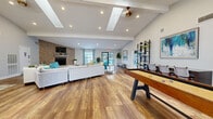 Located in Clearwater, Florida, this single-floor community clubhouse offers a generously sized, thoughtfully designed gathering space for residents. The main lounge area is a standout, featuring warm hardwood-style flooring, vaulted ceilings with exposed beams, skylights, and recessed lighting that floods the space with brightness. A dramatic floor-to-ceiling stacked stone accent wall anchors the room, housing a fireplace topped with a wall-mounted TV, creating an inviting focal point. Comfortable seating arrangements — including vibrant teal accent chairs and neutral sofas — are complemented by a shuffleboard table, making the space ideal for both relaxation and recreation. A direct glass door provides convenient access to an outdoor pool area. The fitness center is well-equipped with treadmills, an elliptical, a cable weight machine, and additional cardio equipment, all set against bold teal accent walls and durable tile flooring. A dedicated business or study room offers built-in dark cabinetry, a long countertop workspace, and a small seating area, providing a quiet and functional environment. Together, these spaces combine modern coastal design with practical amenities, creating a vibrant and versatile community hub.