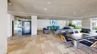 Located at 16101 Murphy Ave in Irvine, CA, this generously sized amenity space delivers a resort-like experience across a single level of thoughtfully designed indoor and outdoor areas. At the heart of the outdoor space is a large, sparkling swimming pool framed by wide concrete decking, lined with chaise lounges and shaded by towering palm trees — an ideal setting for relaxation under the Southern California sun. Adjacent to the pool, a separate spa offers a more intimate retreat, accented by decorative mosaic tilework and lush greenery.

The covered indoor-outdoor clubroom provides a seamless transition between interior comfort and the open-air environment. Light-toned tile flooring, structural columns, and a textured ceiling define the space, which is furnished with wicker lounge seating upholstered in navy and teal, a pool table, and a flat-screen display. A dedicated dining and kitchen area features built-in counter stations with sinks, ample table seating, and recessed lighting, making it well-suited for gatherings and events.

Outdoor pathways wind through manicured landscaping with native plantings, ornamental grasses, and sculptural metalwork gates. Cozy seating clusters with blue cushioned chairs are positioned throughout the grounds, offering inviting spots to unwind. The surrounding multi-story building architecture complements the amenity area with a modern, upscale aesthetic.