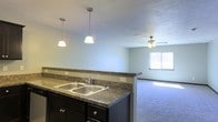 This single-floor space is designed with an open-concept layout that connects the kitchen and living area, creating a cohesive and functional environment. The kitchen features dark espresso-toned cabinetry paired with granite-style countertops that extend into a raised breakfast bar, offering both workspace and a natural divider between the two areas. A double stainless steel sink sits beneath stylish pendant lighting, with two hanging fixtures providing focused illumination over the counter. The living area extends beyond the kitchen, finished with wall-to-wall carpet that adds warmth and comfort underfoot. A ceiling fan centered in the living space provides both air circulation and supplemental lighting. A window along the far wall allows natural light to fill the room, brightening the soft, neutral-toned walls throughout. The open flow between the kitchen and living space makes the layout feel spacious and inviting, while the consistent color palette of cool grays and dark accents lends the space a clean, modern aesthetic. The single-floor design ensures easy accessibility and a straightforward, practical layout throughout.