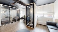 Located at 12-24 Leith Hill Road in Toronto, Ontario, this single-floor office space delivers a refined, contemporary aesthetic across a compact but well-utilized footprint. The space comprises a main living/lounge area and a connecting hallway, thoughtfully designed to balance open collaboration with private functionality.

The lounge area is an industrial-modern blend, anchored by an exposed concrete ceiling with visible ductwork and conduit that adds an urban, loft-like character. Warm wood-tone flooring runs throughout, softening the industrial elements overhead. A standout feature is the built-in media and fireplace console — a sleek, low-profile unit clad in warm wood with an integrated linear electric fireplace and a centrally mounted flat-screen display, flanked by decorative plants and accent objects.

Seating includes a full-length charcoal leather sofa paired with a round, low-profile coffee table, creating a relaxed yet professional lounge setting. A wall-mounted split air conditioning unit ensures year-round climate control.

A prominent freestanding privacy pod — a self-contained acoustic booth with glass door and built-in desk seating — provides a dedicated space for focused work or private calls. Adjacent to it, a glass-partitioned enclave offers additional semi-private seating. Wall sconces provide warm ambient lighting throughout, complementing the natural light from nearby windows.