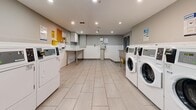 Located at 24 Forest Manor Rd in Toronto's north end, this communal laundry facility offers a clean, efficiently organized space designed to serve building residents. The room is finished with light-toned large-format ceramic floor tiles and white tile wainscoting along the lower walls, contributing to a bright and hygienic environment. Recessed ceiling lighting illuminates the space evenly throughout.

Multiple front-loading washing machines are arranged along one wall, complemented by a full row of top-loading dryers on the opposite side, providing ample capacity for building residents. A large stainless steel utility sink is positioned near the entry door, offering a convenient area for hand-washing or pre-treating garments. A fold-out countertop surface provides space for sorting and folding laundry.

The room is accessed through a solid metal door equipped with a hydraulic door closer, ensuring secure and controlled entry. Informational signage and usage instructions are posted throughout the space, and a fire extinguisher is mounted near the entrance for safety compliance. The overall design is practical and no-frills, prioritizing functionality and ease of use in a well-maintained shared amenity space.