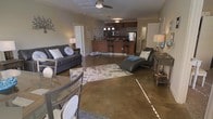 This single-floor residence showcases an open-concept layout that seamlessly connects the living, dining, and kitchen areas into one cohesive, flowing space. The polished concrete floors throughout lend an industrial-modern aesthetic, reflecting light and contributing to the spacious feel of the interior.

The living area is anchored by a gray sofa adorned with decorative pillows, complemented by a leather chaise lounge and a patterned area rug. A ceiling fan overhead provides both function and a subtle design element. The dining area, positioned toward the front, features a glass-top table with contemporary seating.

The kitchen is a standout feature, boasting dark cabinetry with warm wood tones, stainless steel appliances including a refrigerator and microwave, and a raised breakfast bar with bar stools — ideal for casual dining or entertaining. A pendant light fixture above the bar adds a refined touch.

Neutral wall tones throughout create a calm, cohesive backdrop, while accent lighting from floor lamps adds warmth. The layout maximizes functionality while maintaining an open, airy ambiance across the entire space.