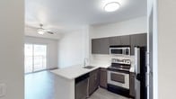 This single-floor residence showcases a well-designed open-concept layout where the kitchen flows seamlessly into the living area. The kitchen is a standout feature, equipped with stainless steel appliances including a range, microwave, and refrigerator. Dark espresso-toned cabinetry provides generous storage both above and below the countertops, contrasting attractively with the light-colored countertop surfaces. A peninsula with an undermount sink and built-in dishwasher extends into the space, offering additional prep area and a natural dividing point between the kitchen and living zone. The flooring transitions between wood-look planks in the kitchen and living areas, adding warmth and continuity throughout. The living area benefits from a ceiling fan with light fixture, contributing to both comfort and illumination. A sliding glass door floods the space with natural light and provides direct access to a private balcony, enhancing the overall sense of openness. The textured ceiling and neutral wall tones create a clean, contemporary backdrop throughout the unit.