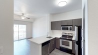 This single-floor residence showcases a well-designed open-concept layout where the kitchen flows seamlessly into the living area. The kitchen is a standout feature, equipped with stainless steel appliances including a range, microwave, and refrigerator. Dark espresso-toned cabinetry provides generous storage both above and below the countertops, contrasting attractively with the light-colored countertop surfaces. A peninsula with an undermount sink and built-in dishwasher extends into the space, offering additional prep area and a natural dividing point between the kitchen and living zone. The flooring transitions between wood-look planks in the kitchen and living areas, adding warmth and continuity throughout. The living area benefits from a ceiling fan with light fixture, contributing to both comfort and illumination. A sliding glass door floods the space with natural light and provides direct access to a private balcony, enhancing the overall sense of openness. The textured ceiling and neutral wall tones create a clean, contemporary backdrop throughout the unit.