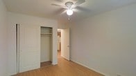 This single-floor residence offers a clean, functional layout with a warm and inviting aesthetic. The bedroom is a standout feature, showcasing light-toned hardwood-style flooring that extends throughout the space, contributing to a cohesive and airy feel. A ceiling fan with an integrated light fixture is centrally mounted, providing both comfort and illumination. The bedroom includes a built-in closet with a hanging rod, offering practical storage in a compact, well-organized space. Neutral white walls keep the interior feeling bright and open, while white panel doors complement the overall clean design. A doorway from the bedroom leads directly to the rest of the home, with a glimpse into what appears to be a kitchen area featuring cabinetry and appliances visible in the background. The simple, understated design of the space makes it easy to personalize, and the consistent flooring and neutral palette create a seamless flow between rooms. Overall, the residence presents a tidy, functional living environment suited to a variety of lifestyles.