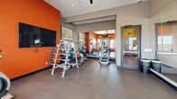 Located at 301 Nichols Street in Fort Worth, Texas, this fitness center is a thoughtfully designed exercise space that packs a full range of workout options into a compact, single-floor layout. The room immediately draws attention with its bold orange accent walls, which lend a high-energy atmosphere complemented by neutral gray tones on the surrounding walls and polished concrete flooring throughout.

Large windows fitted with horizontal blinds line one side of the room, allowing natural light to pour in while offering views of the surrounding Fort Worth skyline. A ceiling fan ensures comfortable air circulation during workouts. Full-length mirrors along one wall visually expand the space and provide functional feedback for exercise form.

The fitness center is well-appointed with a diverse selection of equipment. Cardio options include treadmills, an elliptical trainer, and a rowing machine. A dedicated free weights area features a dumbbell rack with a variety of weights, an adjustable workout bench, and stability balls. A multi-function cable and strength machine rounds out the strength training offerings. Additional accessories such as resistance bands and small fitness tools are neatly organized on a utility rack.

Decorative framed artwork adds a polished touch, reinforcing the space's clean and motivating aesthetic. The fitness center is accessed through a glass-panel door, providing a clear sightline into the room from the entrance.