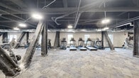 Located at 611 S Wright St in Champaign, Illinois, this fitness center offers a comprehensive workout facility spread across a single, generously proportioned floor. The space is defined by its industrial aesthetic, featuring an exposed dark ceiling with visible ductwork, piping, and structural beams, all complemented by bright overhead lighting that ensures clear visibility throughout.

The cardio zone is well-equipped with multiple treadmills and elliptical trainers arranged in an organized row, providing ample space between each machine. A dedicated free weights area includes a full rack of dumbbells, a weight bench, and barbell equipment, suited for strength training of all levels. Several selectorized cable and resistance machines are distributed across additional sections of the floor, catering to targeted muscle group workouts.

A distinct flexibility and functional training area features yoga mats, stability balls, medicine balls, and a water cooler station, anchored by a bold star logo graphic on the wall. The flooring transitions between patterned carpet tiles and rubber matting in heavier lifting zones, offering both comfort and durability. Natural light filters in through perimeter windows, and the open layout, supported by prominent wrapped structural columns, creates an industrial yet functional training environment.