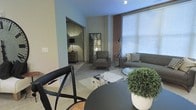 This single-floor space offers a well-designed open layout that connects the dining and living areas in a cohesive, functional arrangement. The dining area is positioned at the forefront, furnished with a sleek dark round table and matching chairs, anchoring the space with a contemporary feel. A lush potted plant adds a fresh, organic accent to the setting.

The living area extends naturally from the dining space, centered around a comfortable gray sofa and matching armchair arranged on a cowhide-patterned area rug. A standout architectural feature is the exposed brick accent wall, which introduces texture and warmth against the otherwise neutral, light-toned walls. Large windows fitted with horizontal blinds allow generous natural light to fill the room while maintaining privacy.

An adjoining room, visible through an open doorway, provides additional space and is accented by a vintage-style mirror and a small wood dresser with a table lamp, suggesting a bedroom or flexible secondary space. Track lighting overhead complements the natural light throughout. The overall ambiance is modern yet warm, blending industrial and contemporary design elements cohesively.
