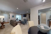This thoughtfully designed single-floor residence offers a seamless, open layout connecting its primary living spaces. The living room serves as the heart of the home, anchored by a tiled fireplace with a classic white mantel and a wall-mounted television framed by built-in shelving. Crown molding and recessed lighting lend a polished finish to the neutral-toned walls and plush carpeted floors throughout. A pass-through opening connects the living area to the galley-style kitchen, which is outfitted with warm wood cabinetry, stainless steel appliances including a gas range and refrigerator, and light tile flooring. The adjacent dining area accommodates a full table and chairs beneath a track lighting fixture. A dedicated entry foyer provides a welcoming transition into the living space. The primary bedroom features sliding glass doors that invite natural light and outdoor views, complemented by soft carpeting and ample dresser space. A second bedroom or office space benefits from a large window arrangement with sheer curtains, filling the room with natural light. The bathroom offers a clean, functional design with a wood vanity, tile flooring, and a shower-tub combination. Throughout the residence, a cohesive neutral palette and consistent finishes create a calm, comfortable ambiance.
