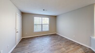 Located at 2404 Hill St in Raleigh, NC, this two-bedroom, one-bathroom residence offers a functional and well-organized single-floor layout. The combined kitchen and living room serves as the heart of the home, featuring wood-look plank flooring, a textured ceiling, and a peninsula counter with granite-style surfaces that separates the kitchen from the living area. The kitchen is equipped with warm wood cabinetry, matching granite-style countertops, black appliances including a refrigerator, range, and dishwasher, and ample cabinet storage on both sides of the galley-style workspace. Natural light filters into the living area through a large window with blinds. Both bedrooms are carpeted and finished in neutral tones, each featuring windows that bring in outdoor light and views of surrounding greenery. The primary bedroom includes direct access to a private balcony with wood decking. The full bathroom is finished with a white tub-shower combination, wood-look flooring, and a vanity with wood cabinetry. A hallway connects the main living spaces, providing a practical flow throughout the unit. The overall aesthetic is clean and neutral, offering a comfortable, move-in-ready living environment.