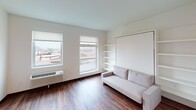 This thoughtfully designed studio unit maximizes functionality across a single floor. The main living area features rich hardwood-style flooring, crisp white walls, and large windows that invite abundant natural light while offering views of the surrounding cityscape. A built-in Murphy bed system with integrated open shelving and a neutral-toned sofa demonstrates smart space-saving design, allowing the room to serve as both a comfortable living and sleeping space.

The kitchen is outfitted with a full suite of stainless steel appliances, including a refrigerator, electric range, and over-the-range microwave. White upper cabinetry contrasts with dark lower cabinets and a dark countertop, creating a modern, two-toned aesthetic. The layout provides efficient workflow despite the compact footprint.

The bathroom is designed with accessibility in mind, featuring grab bars, a wide layout, and a soaking tub alongside the toilet and vanity. Adjacent to the bathroom is a dedicated laundry area with a stacked front-loading washer and dryer unit, adding a high level of convenience rarely found in similarly sized spaces.

Throughout the unit, the consistent hardwood-style flooring, neutral color palette, and clean-lined finishes create a cohesive, modern ambiance that is both practical and welcoming.