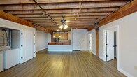 This one-bedroom, one-bathroom unit blends historic industrial character with modern conveniences. The defining architectural feature is the open, exposed wood beam ceiling that spans the entire unit, lending a raw, loft-like aesthetic while adding warmth and visual depth overhead. Ceiling fans are mounted throughout, providing comfortable airflow in each room.

The main living area is generously proportioned, with durable hardwood-style flooring and exposed brick accent walls that reinforce the unit's historic charm. Natural light enters through double-pane windows fitted with blinds. A dedicated in-unit laundry area with a full washer and dryer is conveniently integrated into the living space.

The kitchen features warm wood cabinetry, granite countertops, and a full suite of stainless steel appliances, including a refrigerator, electric range, and over-the-range microwave. A raised breakfast bar provides casual dining or prep space.

The bedroom offers a spacious layout with continued exposed beam ceilings, brick accents, and a wall-mounted mini-split HVAC unit for individualized climate control. The bathroom is finished with light tile flooring, a wood vanity, a soaking tub, and a separate tiled shower with matte black fixtures, offering a clean, modern contrast to the unit's rustic elements.