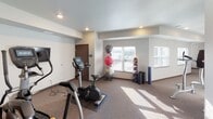 This single-level fitness center offers a comprehensive workout environment designed to accommodate a variety of exercise routines. The space is laid out across an open, L-shaped floor plan with neutral-toned walls, carpeted flooring, and recessed lighting that creates a clean, functional atmosphere throughout.

The cardio zone is equipped with a treadmill, stationary bike, and elliptical machine, positioned to take advantage of natural light streaming in through multiple windows that offer views of the surrounding area. The weight training section includes a multi-tiered dumbbell rack stocked with a full range of free weights, an adjustable workout bench, and a cable weight machine, providing ample options for strength training. A dedicated area for functional fitness features stability balls of varying sizes, a medicine ball rack, and a yoga mat, supporting flexibility and core training needs.

Motivational canvas artwork is displayed throughout, adding visual interest to the otherwise minimalist décor. The space is accessed through a solid door with a glass panel insert. Recessed ceiling lights and a ventilation system ensure a comfortable environment, while the carpeted floors provide cushioning underfoot during workouts.