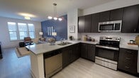 This single-floor residence offers a thoughtfully designed open-concept layout that seamlessly connects the kitchen and living areas. The kitchen is a standout feature, showcasing rich dark cabinetry that contrasts beautifully with light granite countertops. Stainless steel appliances — including a range, microwave, and dishwasher — provide a sleek, modern finish. A peninsula with an undermount sink and pendant lighting above adds both functionality and visual appeal, making it an ideal space for cooking and entertaining. Light wood-tone flooring runs throughout, lending warmth and continuity to the space. The adjacent living area is anchored by a bold blue accent wall that adds a pop of color and personality to the room. Natural light filters in through a window, brightening the space and creating an inviting ambiance. The open flow between the kitchen and living room fosters a connected, social atmosphere, making the space feel expansive and welcoming. Overall, the residence combines contemporary design elements with practical finishes, resulting in a comfortable and stylish living environment suited for everyday living.